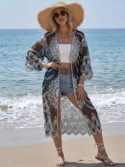 Black-Sheer-Lace-Beach-Kimono-Cover-Up A woman standing on a beach wearing a long sheer black patterned kimono with white lace trim, a white crop top, denim shorts, a straw hat, and strappy sandals.